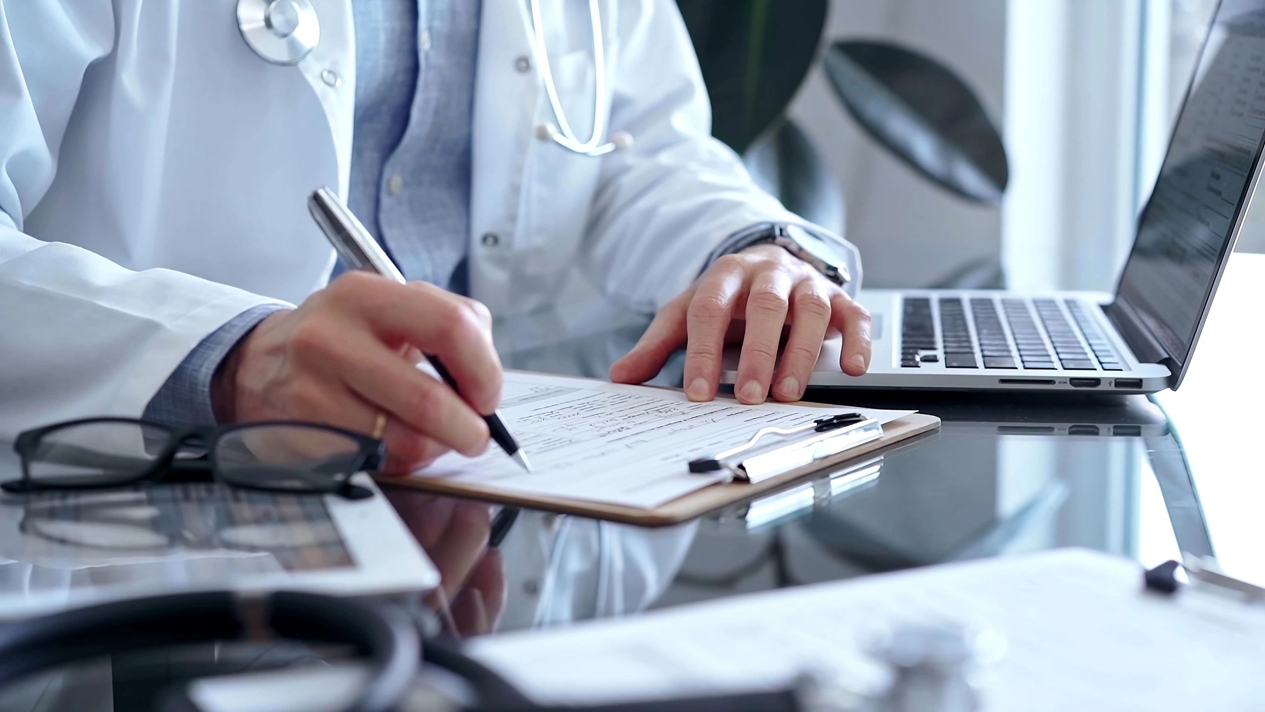 Healthcare professional writing on clipboard with laptop